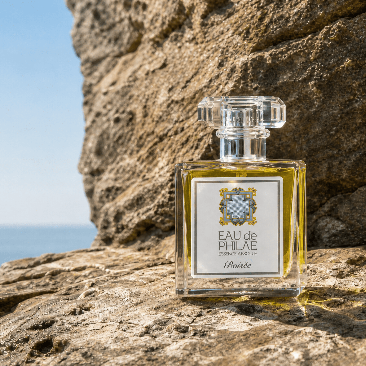 A square glass perfume bottle placed on a rocky surface outdoors with a blue sky and sea in the background. Eau De Phile Boisee organic perfume for men, sold by Inlight Beauty
