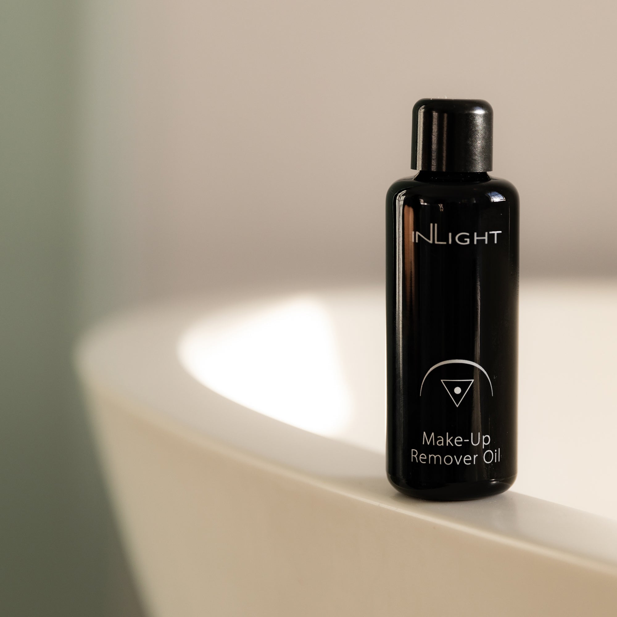A small black bottle of organic Make-Up Remover Oil standing on the rim of a white bathtub in a softly lit bathroom setting. Inlight Beauty