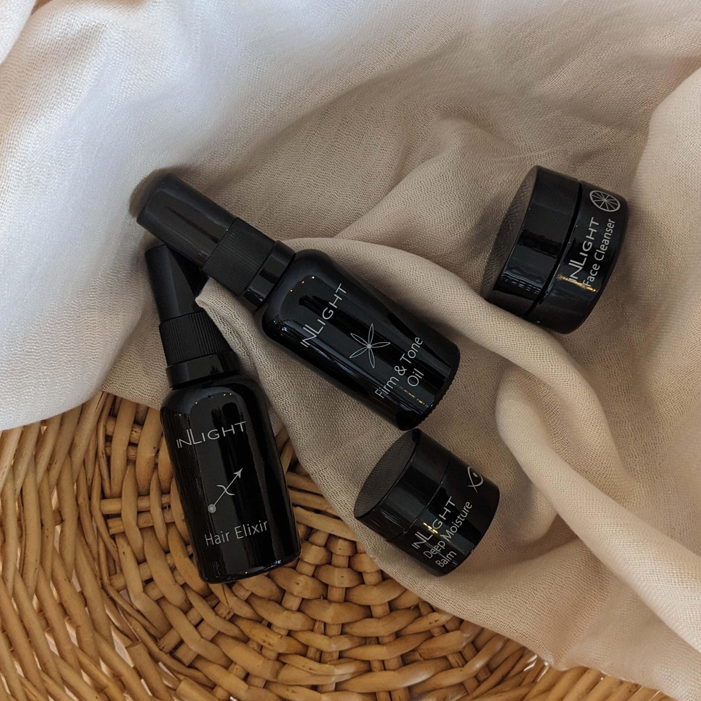Black bottles with white labels on a beige cloth and woven basket. Inlight beauty organic summer skincare. Body oil for toning. Organic cleansing balm.