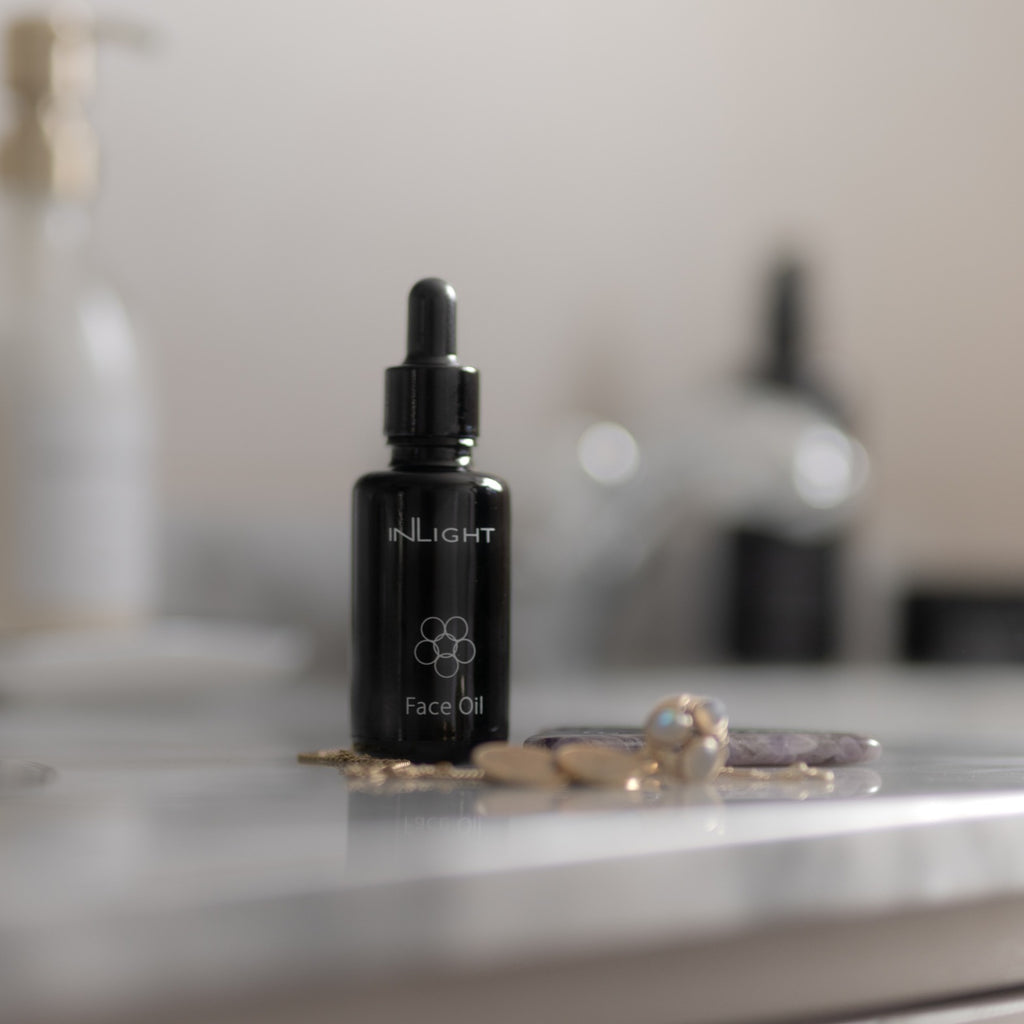 A small black dropper bottle labeled INLIGHT Face Oil sits on a light marble surface with blurred toiletries in the background.